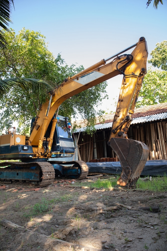 Excavatrice Kobelco puissante : travail de terrassement en extérieur. Excavatrice Kobelco jaune et bleue sur chenilles, la flèche levée et le godet au sol, devant un hangar en bois et des arbres verts.