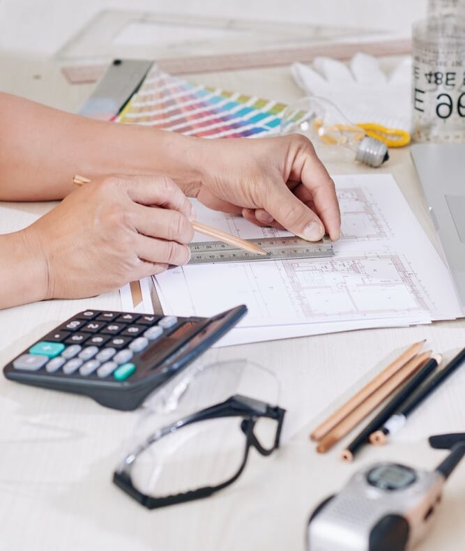 Architecte en action : Mesurer un plan de construction précis. Architecte mesurant un plan de construction avec règle et crayon. Sur le bureau : calculatrice, lunettes de sécurité et nuancier de couleurs.