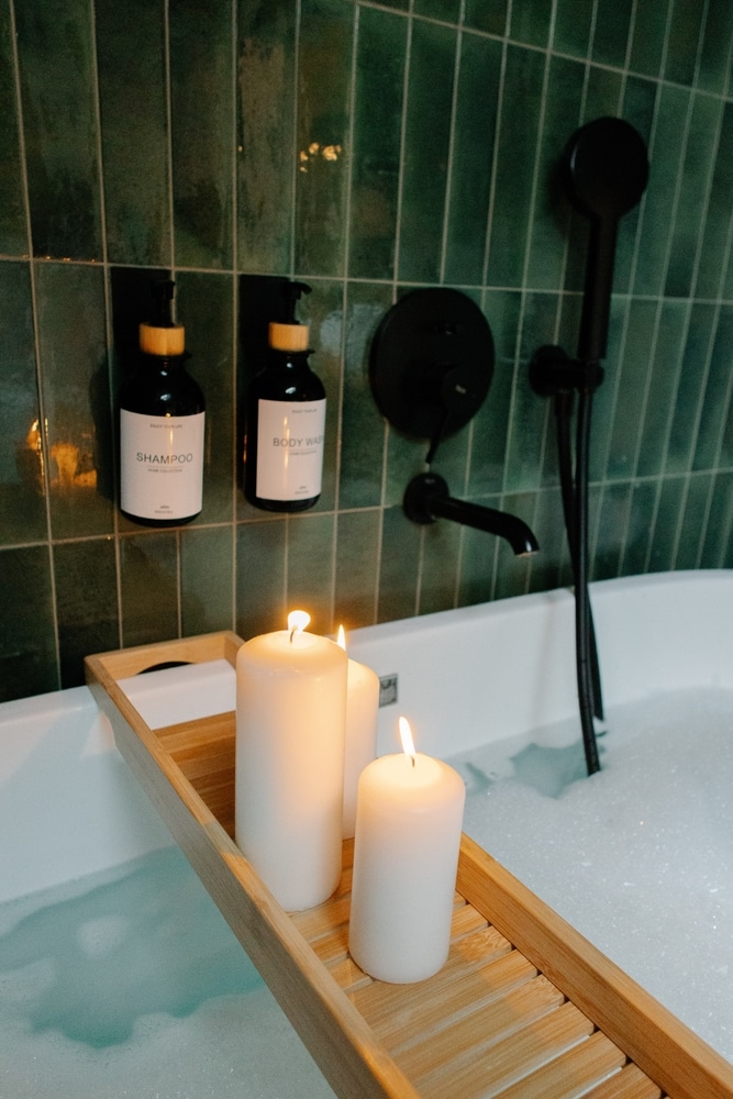 Bain détente luxe : bougies, carreaux verts et ambiance spa. Bain relaxant aux chandelles. Carreaux verts, robinetterie noire mate. Plateau en bambou avec bougies. Produits SHAMPOO et BODY WASH fixés au mur.