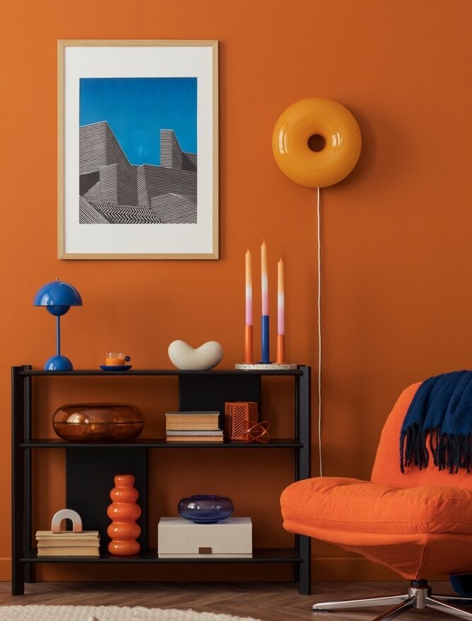 Salon moderne : idées déco orange terracotta et bleu vif. Décoration moderne audacieuse : mur orange, étagère noire, fauteuil orange, lampe bleue et œuvre d'art graphique encadrée.