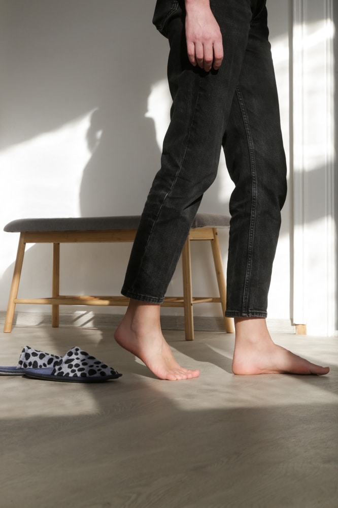 Pieds nus en jeans noirs: confort et style à la maison. Pieds nus et jeans noirs sur sol clair, s'éloignant de chaussons tachetés près d'une banquette. Forte lumière naturelle et ombre.